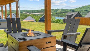 A fire pit with chairs, a wooden deck, and a view of a lake and buildings in the background.