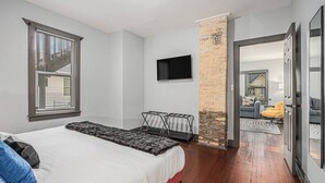 A bedroom with a bed, a TV mounted on the wall, a brick column, and a view into another room through an open door. - Luxe Michigan City Getaway-Private-Unit A (Michigan City)