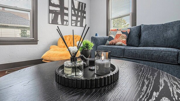 A modern living room with a dark wooden coffee table, a sofa, and a yellow armchair. There are abstract paintings on the wall and a window with blinds. - Luxe Michigan City Getaway-Private-Unit A (Michigan City)
