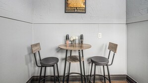 A corner of a room with a white tiled wall, a framed poster of Indiana Dunes National Park, a round wooden table with two chairs, and a small vase with flowers. - Luxe Michigan City Getaway-Private-Unit A (Michigan City)