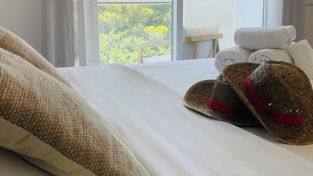 A hotel room with a large window, a bed with white linens, a woven pillow, and a pair of straw hats. - Casa Bonic: Villa with Sunset - seaview and pool (Sant Josep de sa Talaia)