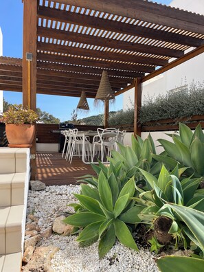 Un patio al aire libre con una pérgola de madera, sillas blancas, una mesa y plantas en macetas.