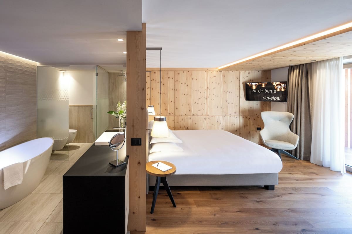 Ein modernes Hotelzimmer mit einem großen Bett, einem separaten Badezimmer mit Badewanne, einem Schreibtisch mit Lampe und einem an der Wand montierten Fernseher.
