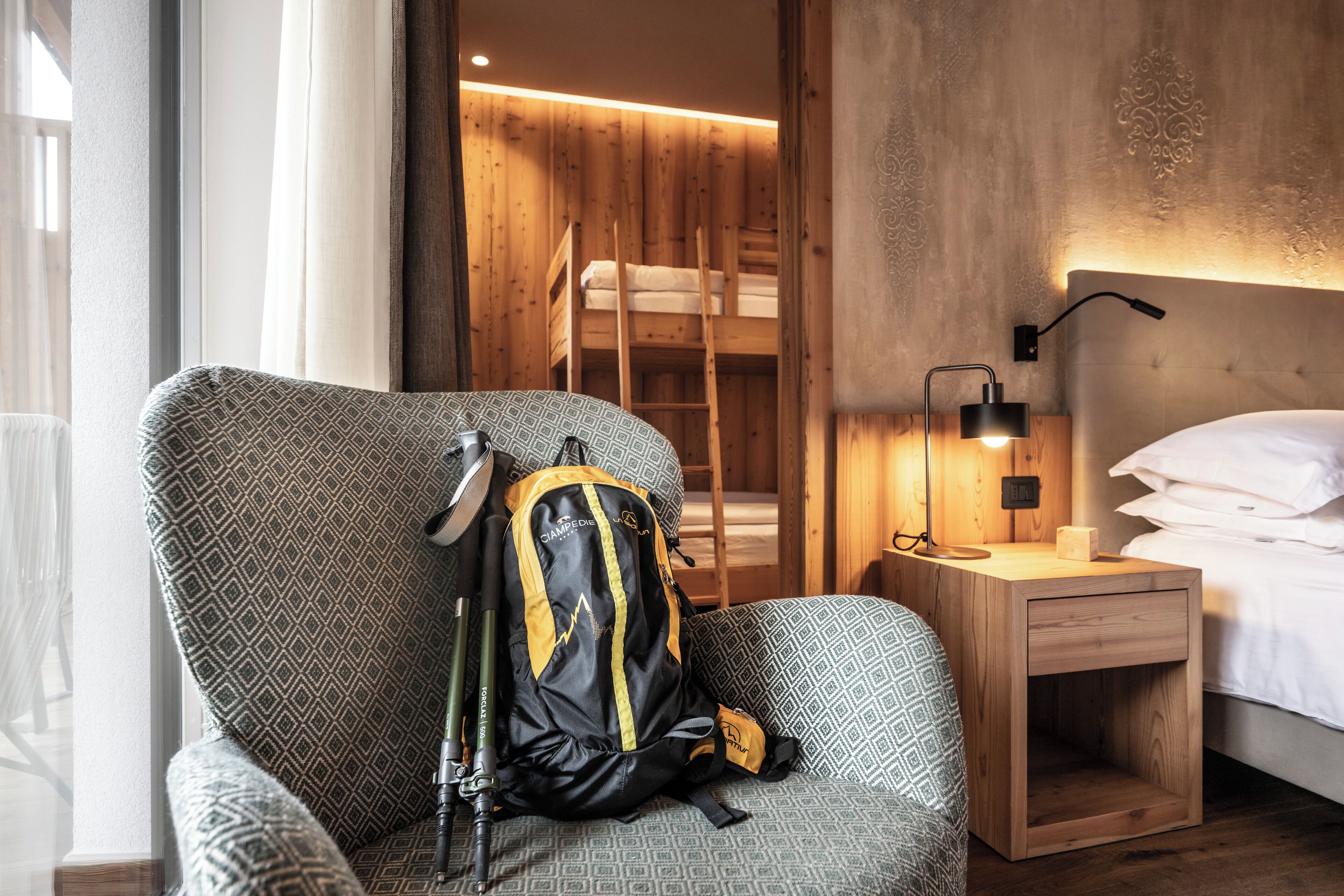 A hotel room with a patterned armchair, a wooden bunk bed, a bedside table with a lamp, and a backpack on the armchair.