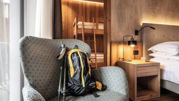 A hotel room with a patterned armchair, a wooden bunk bed, a bedside table with a lamp, and a backpack on the armchair.