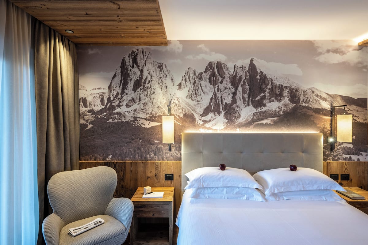 Ein Hotelzimmer mit Bergbild, einem Bett mit weißer Bettwäsche, einem Holznachttisch, einem Stuhl und einem Schreibtisch mit einer Zeitung.