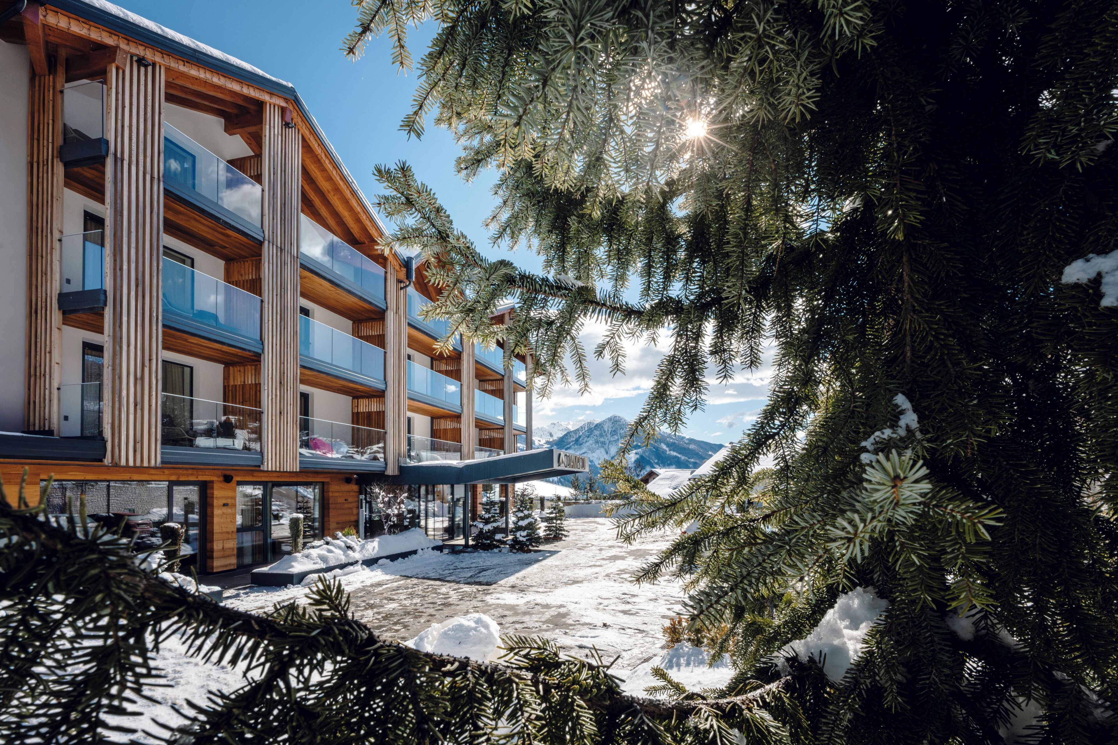 A modern multi-story building with large windows and balconies, surrounded by snow-covered ground and pine trees.