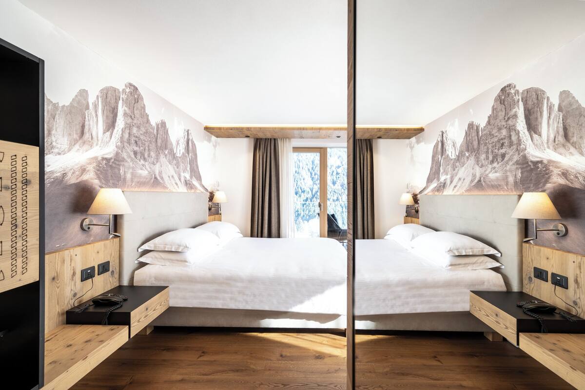 Ein Hotelzimmer mit Spiegel, zwei Betten, Holz-Kopfteilen, Nachttischen mit Lampen und einem großen Wandbild mit Bergmotiv.