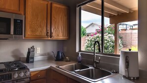 A modern kitchen with wooden cabinets, a stainless steel sink, a microwave, a gas stove, and a window with a view of a garden. - Quiet Escape- Family Getaway by Trails and Shopping (Tucson)
