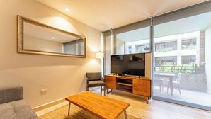Ein modernes Wohnzimmer mit einer Couch, einem hölzernen Couchtisch, einem Fernseher auf einem Holzschrank, einem großen Spiegel und einer Glastür, die zu einem Außenbereich führt.