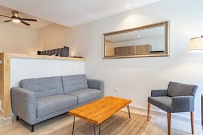 Ein modernes Wohnzimmer mit einem grauen Sofa, einem Holztisch und einem passenden Sessel. An der Wand hängt ein großer Spiegel, und eine Deckenleuchte ist zu sehen.