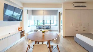 Un salón moderno con una mesa redonda de madera, sillas blancas, un sofá gris con cojines estampados y un televisor montado en la pared.