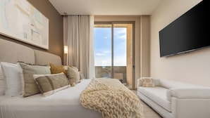 A modern hotel room with a large bed, a sofa, a flat-screen TV, and a balcony with a view.