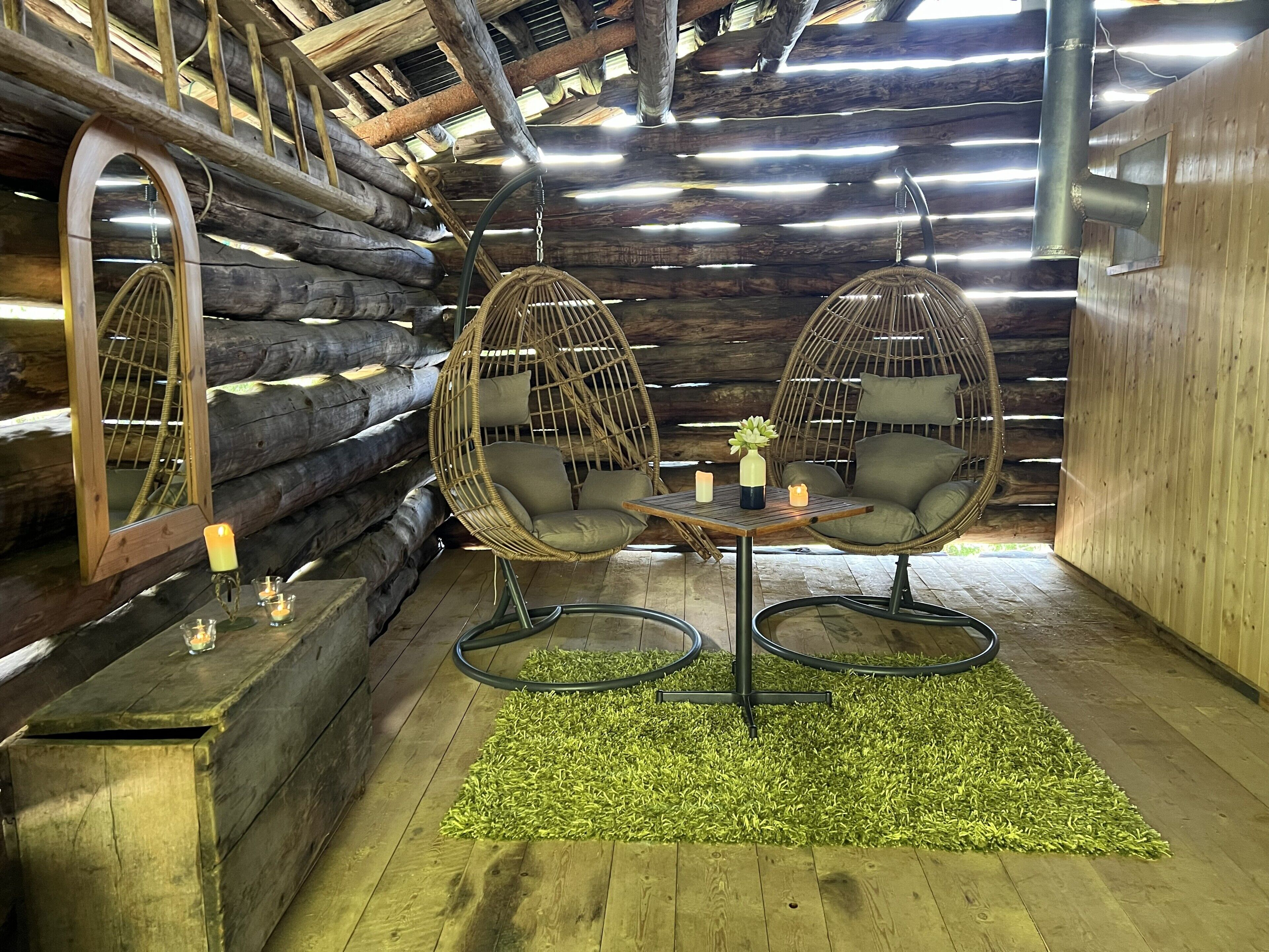 A cozy room with wooden log walls, two hanging chairs, a small table with candles, and a green rug.