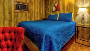 A bedroom with a wooden wall panel, a bed with blue bedding, a red chair, a bedside table with a lamp, and a painting on the wall. - Squirrel Cabin at Panther Creek (Cherokee)
