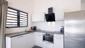 A modern kitchen with white cabinets, a stainless steel refrigerator, a built-in oven, and a window with blinds. - Casa Monrel, Mambo Beach (Willemstad)