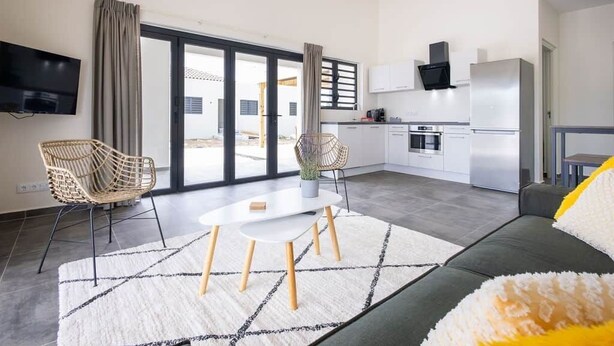 A modern living room with a sofa, a dining table, and chairs, a kitchen area with appliances, and a large sliding glass door leading to an outdoor area. - Casa Monrel, Mambo Beach (Willemstad)