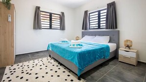 A bedroom with a bed, nightstand, and two windows with curtains. - Casa Monrel, Mambo Beach (Willemstad)