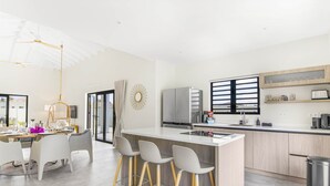 A modern kitchen with a central island, bar stools, a refrigerator, and a window with blinds.