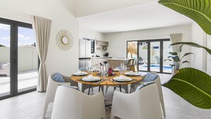 A modern dining area with a round table set for six, surrounded by white chairs. A kitchen is visible in the background, and a large plant is in the foreground.