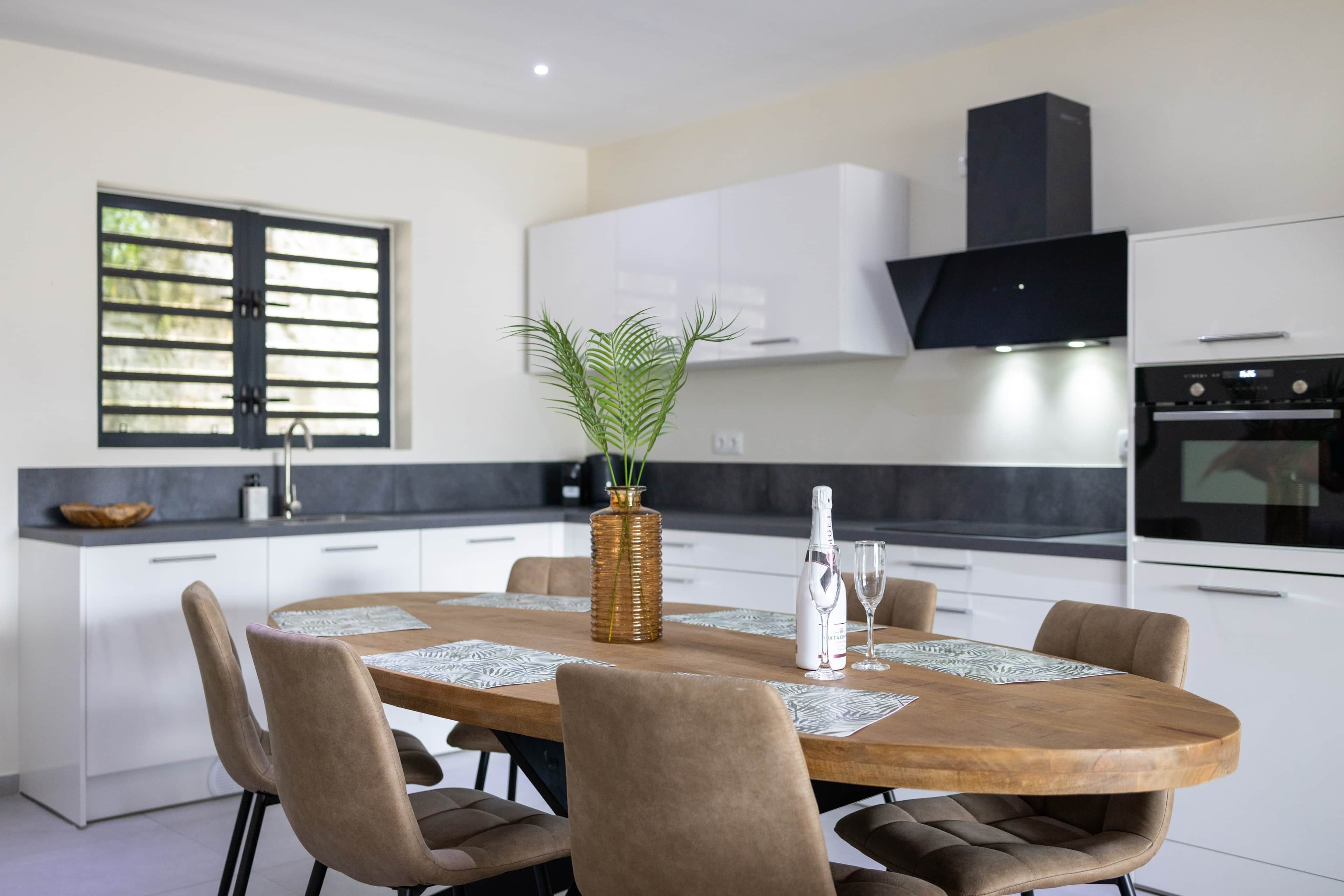 A modern kitchen with a round dining table, chairs, a potted plant, and a wine bottle.