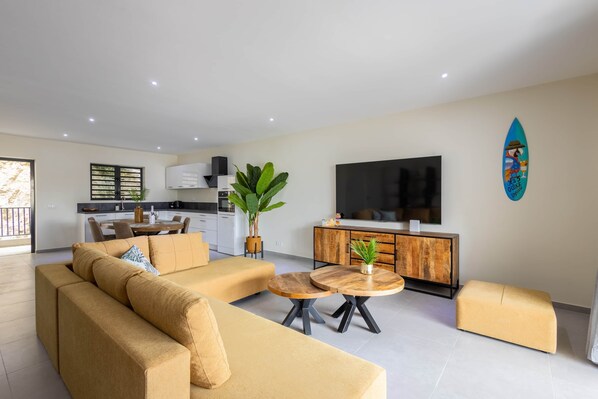 A modern living room with a sofa, ottoman, and round wooden table. There is a kitchen in the background with a dining table and chairs.