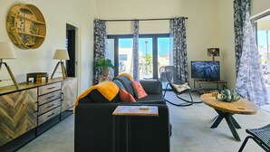 Una sala de estar moderna con un sofá, un televisor, una mesa de centro de madera y vistas al mar a través de amplios ventanales.