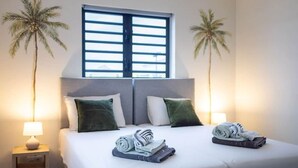 Habitación de hotel con cama, dos almohadas, mesita de noche con lámpara y dos palmeras.