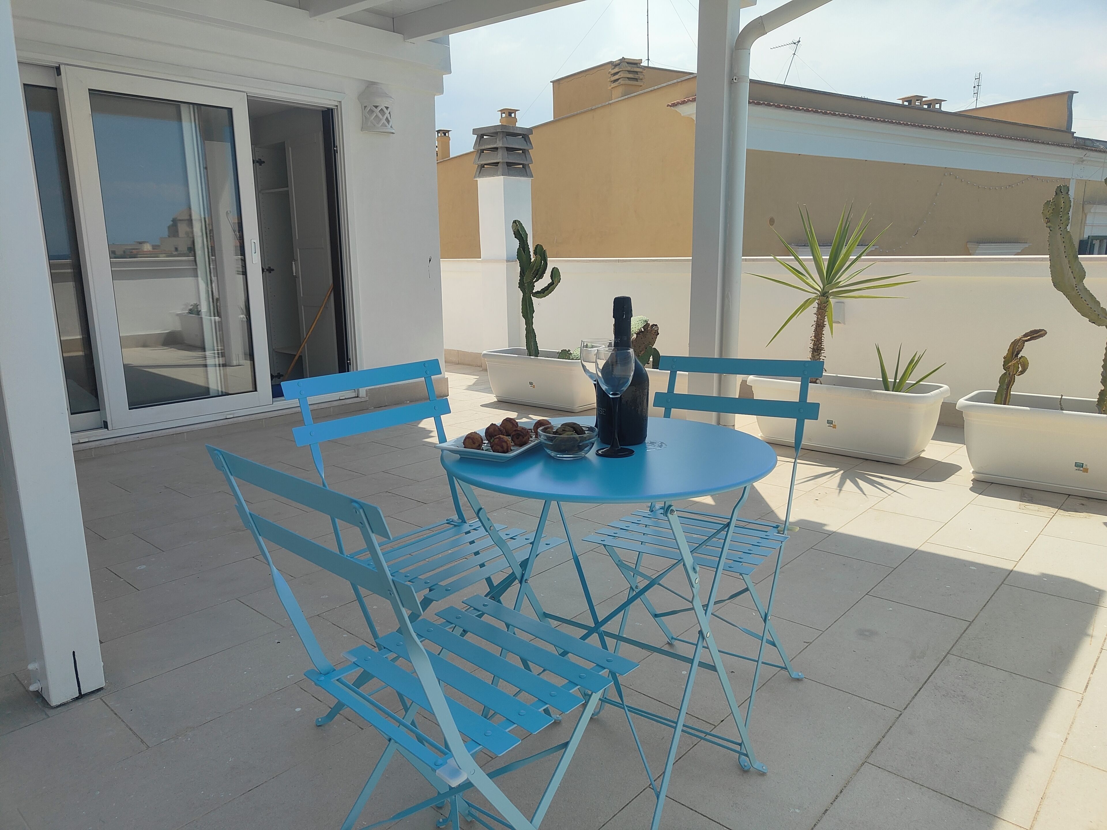 Un conjunto de mesa y sillas de metal azul sobre un patio con baldosas, con una pared blanca y puertas correderas de cristal al fondo.