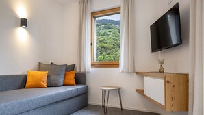 A modern living room with a grey sofa, orange cushions, a small round table, a wooden cabinet, and a TV mounted on the wall. - 305B Apartments No 10 (Ponte Gardena)
