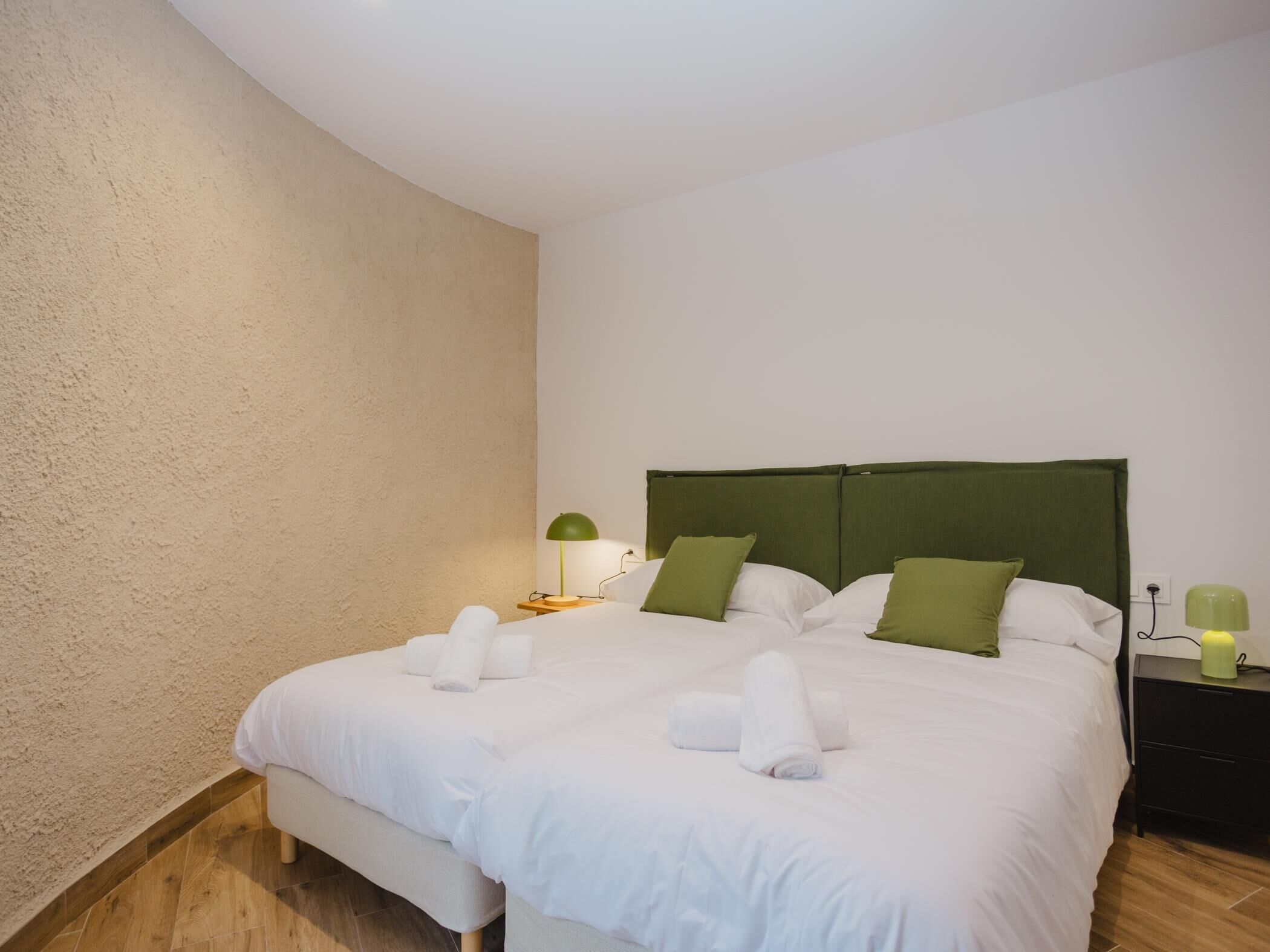 A neatly made double bed with white linens and green pillows, a bedside table with a lamp, and a nightstand with a lamp.