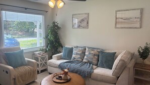 Ein Wohnzimmer mit beiger Couch, gestreifter Dekokissen, einem Couchtisch mit Vase und Fernbedienung sowie einem großen Fenster mit Jalousien.