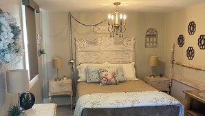 Un dormitorio con cama, mesitas de noche, una araña de techo, un cartel de bienvenida y un detalle decorativo en la pared.