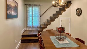 Un comedor con mesa de madera, sillas, candelabro, escalera y un cuadro en la pared.