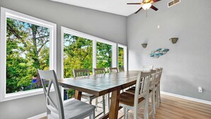 A dining area with a large table and chairs, large windows with a view of trees, a ceiling fan, and wall sconces. - Greens and Gators For 20ppl (Lake Ozark)