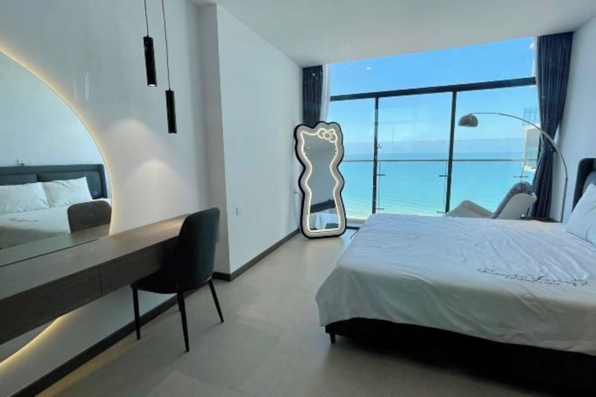 A modern hotel room with a large bed, a desk, a chair, a large window with a view of the sea, and a unique mirror design.
