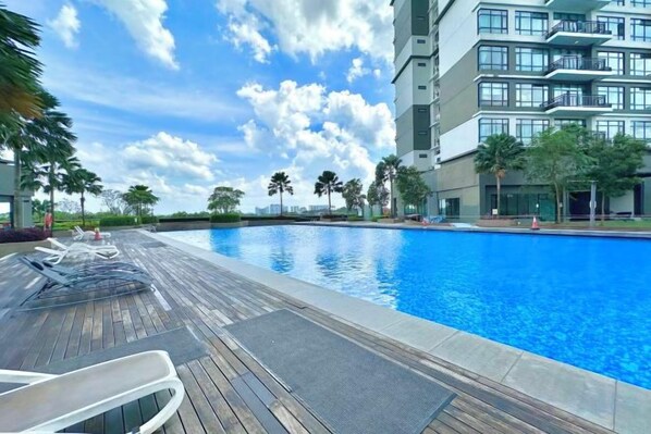 A swimming pool with a wooden deck, lounge chairs, and a modern building in the background. - Molek Regency By Stayrene (Plentong)
