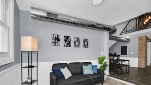 A modern living room with a black leather sofa, a floor lamp, abstract wall art, and a staircase leading to an upper level.