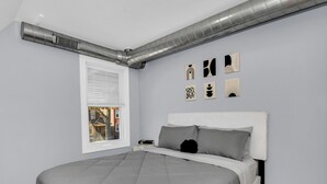 A bedroom with a bed, a window, a wall-mounted air duct, and several wall art pieces.