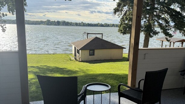 A covered patio with two chairs and a small table, overlooking a grassy area and a building with a sloped roof.
