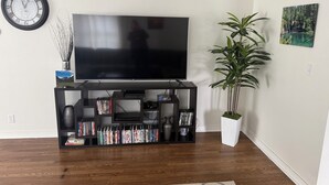 A living room with a flat-screen TV on a black stand, surrounded by bookshelves, a potted plant, and a clock on the wall.