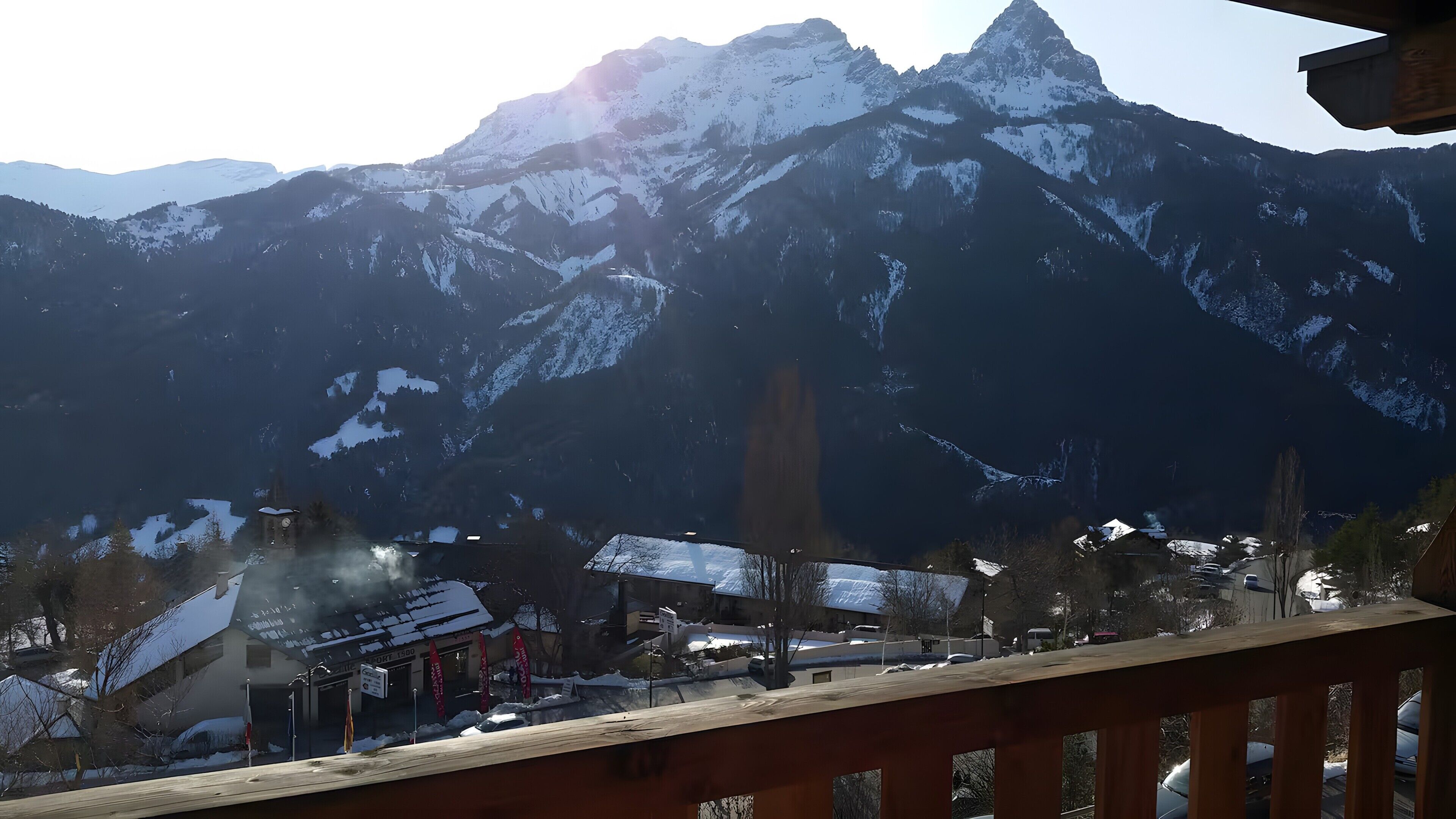 Blick auf Berge mit schneebedeckten Gipfeln, darunter eine Stadt und ein Geländer aus Holz.