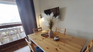 Una mesa de comedor con sillas, un jarrón con flores secas, un tarro de mermelada y un televisor fijado a la pared.