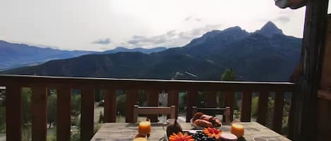 A wooden table with a tray of fruit, a croissant, and a glass of orange juice, overlooking a mountainous landscape.