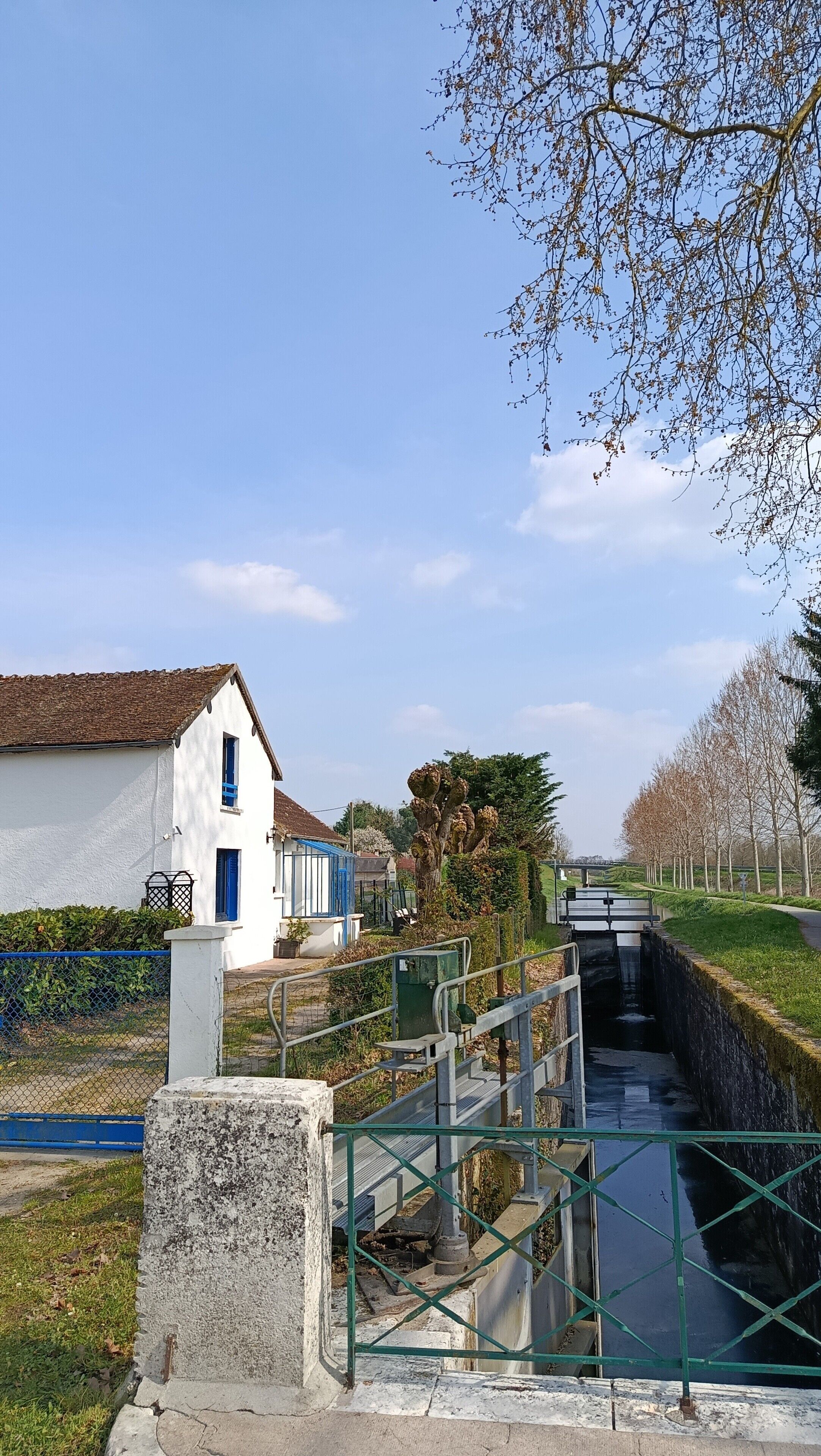 A rural setting with a white building, a narrow water channel, and a tree-lined path.