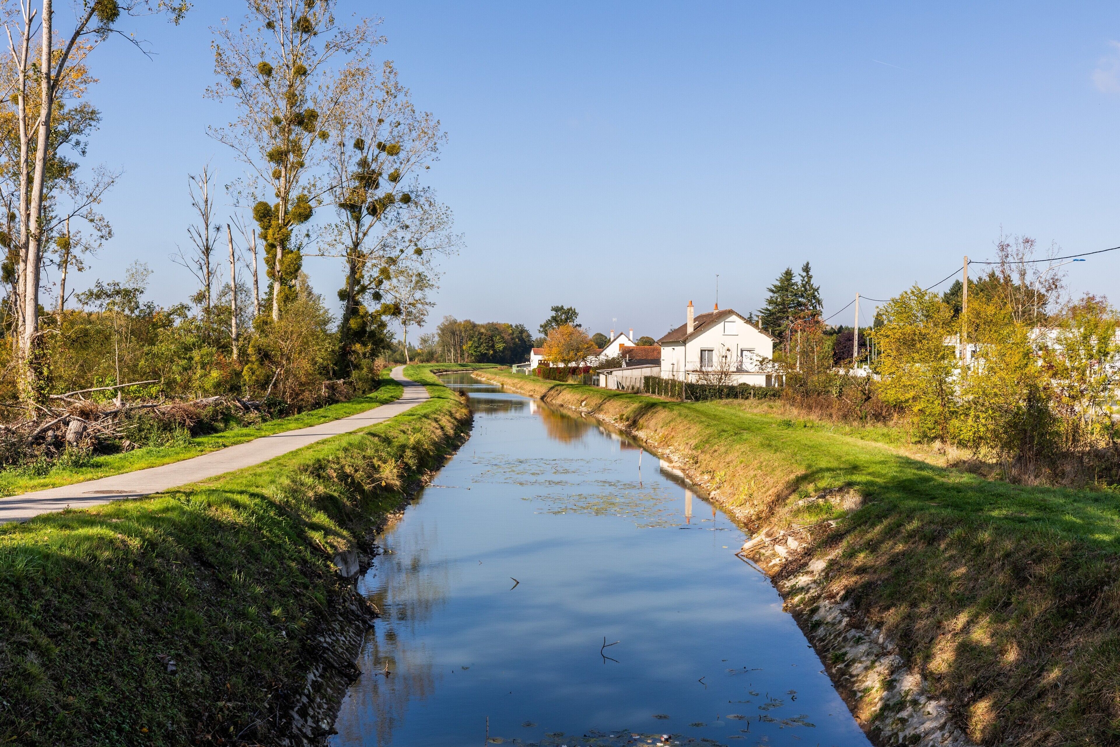 A canal with a pathway on one side, surrounded by greenery and a white building in the distance.