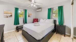 Un dormitorio con cama, ventilador de techo, mesita de noche, cómoda y ventana con cortinas.