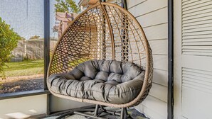 Una silla colgante con asiento y respaldo acolchados, sostenida por una estructura de cuerda trenzada, frente a una fachada de edificio.
