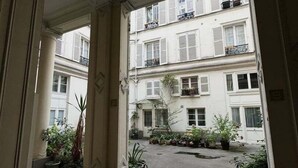 A corridor with a view of a courtyard with potted plants and a building with balconies.