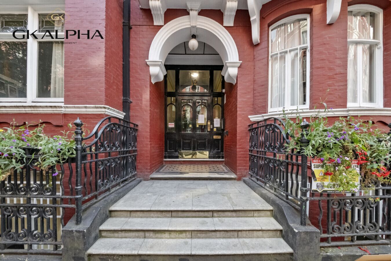 A red brick building with a black front door, steps leading up to the entrance, and black wrought iron railings.